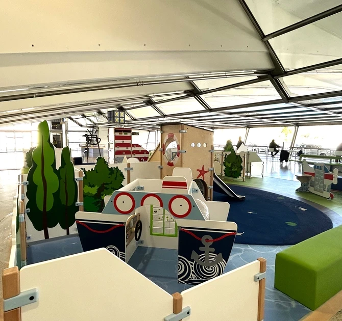 Speelzone op Bilbao Airport met maritiem thema, inclusief speelschip, vuurtoren, glijbaan en vliegtuigje, omgeven door comfortabele zitbanken voor ouders in een lichte vertrekhal.