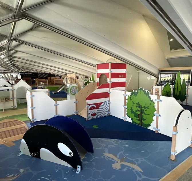 "Overzicht van de kinderspeelhoek op Bilbao Airport, met maritiem thema zoals een vuurtoren, houten roeiboot en walvis-speelelementen, ontworpen voor spel en ontspanning tijdens het wachten."
