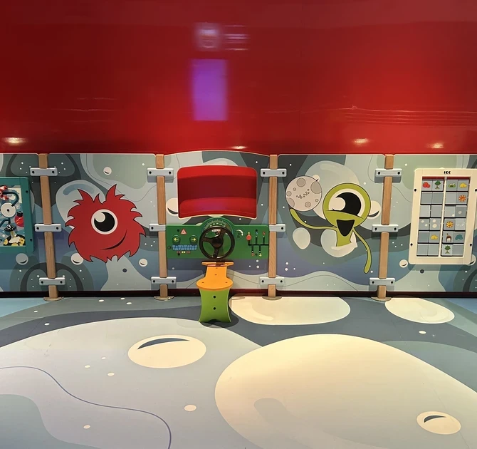 Kinderspeelwand met ruimte-thema, kleurrijke buitenaardse figuren en interactieve spelelementen zoals stuurwiel, schuifpuzzels en spelpanelen, geplaatst tegen een rode wand.