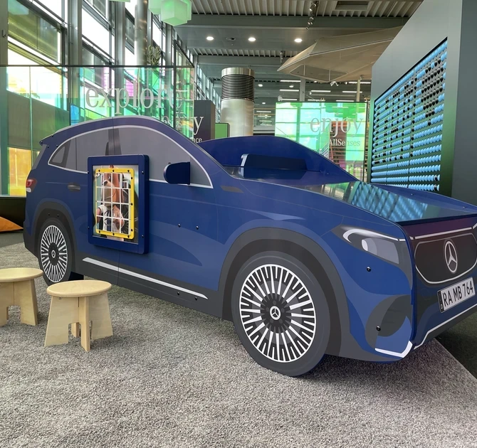 Blauw Mercedes-auto speelmeubel met wandspel, houten krukjes en kleurrijke zitzakken op de achtergrond, geplaatst in de kinderhoek van het Mercedes-Benz Kundencenter in een moderne showroomomgeving.